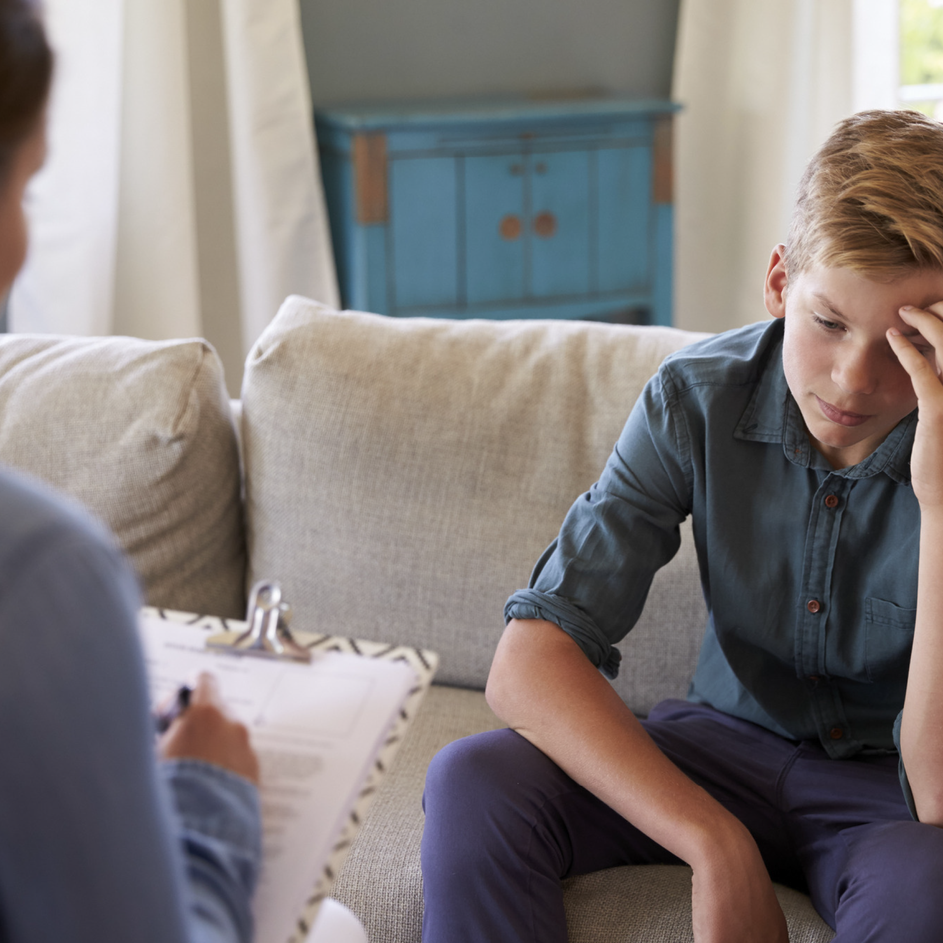 A boy sits on a couch, leaning his forehead on his hand, talking to a woman with a clipboard.
